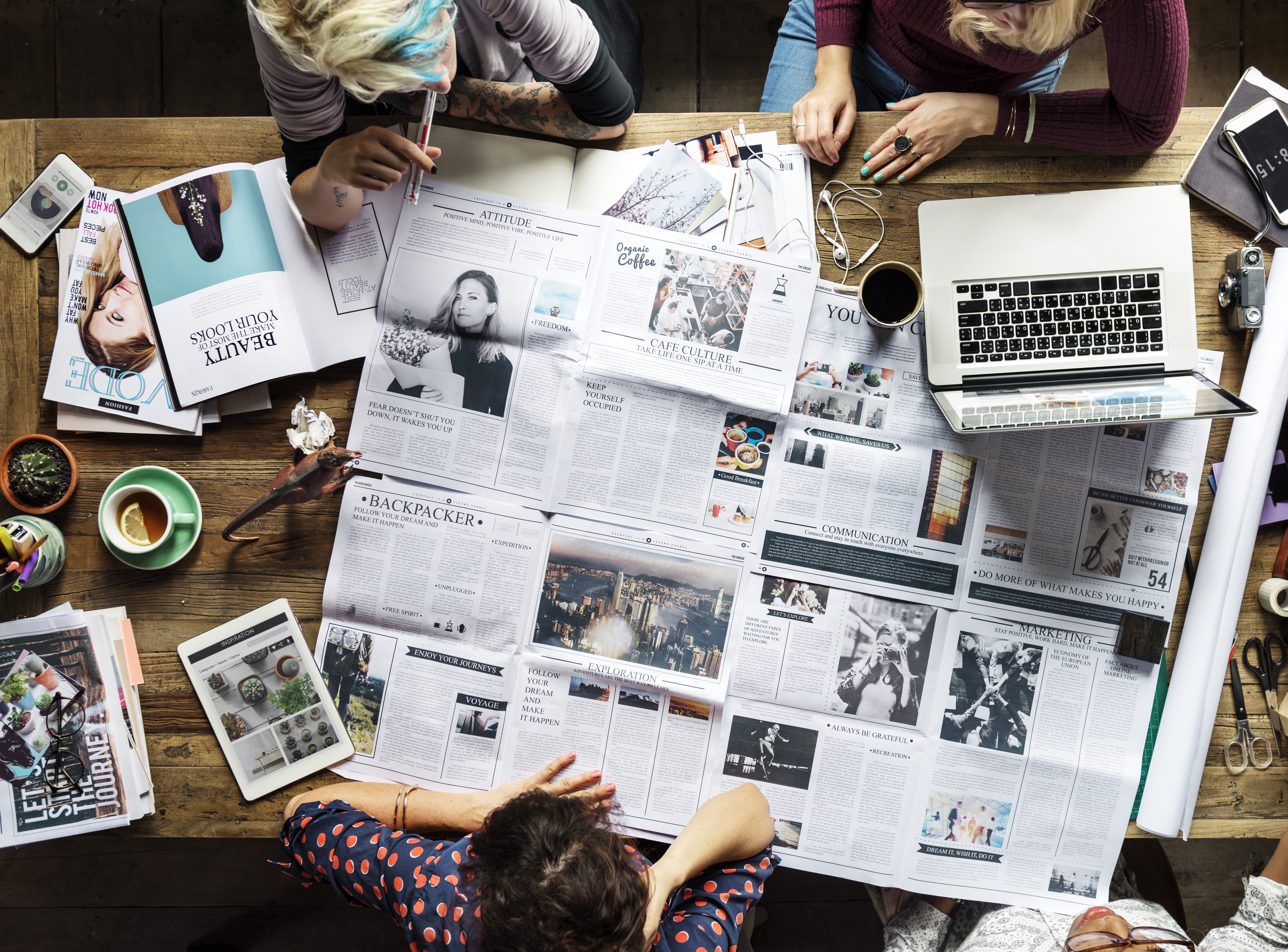 Overhead view of a creative team working together with newspapers, magazines, a laptop, coffee, and design tools spread out on a wooden table.
