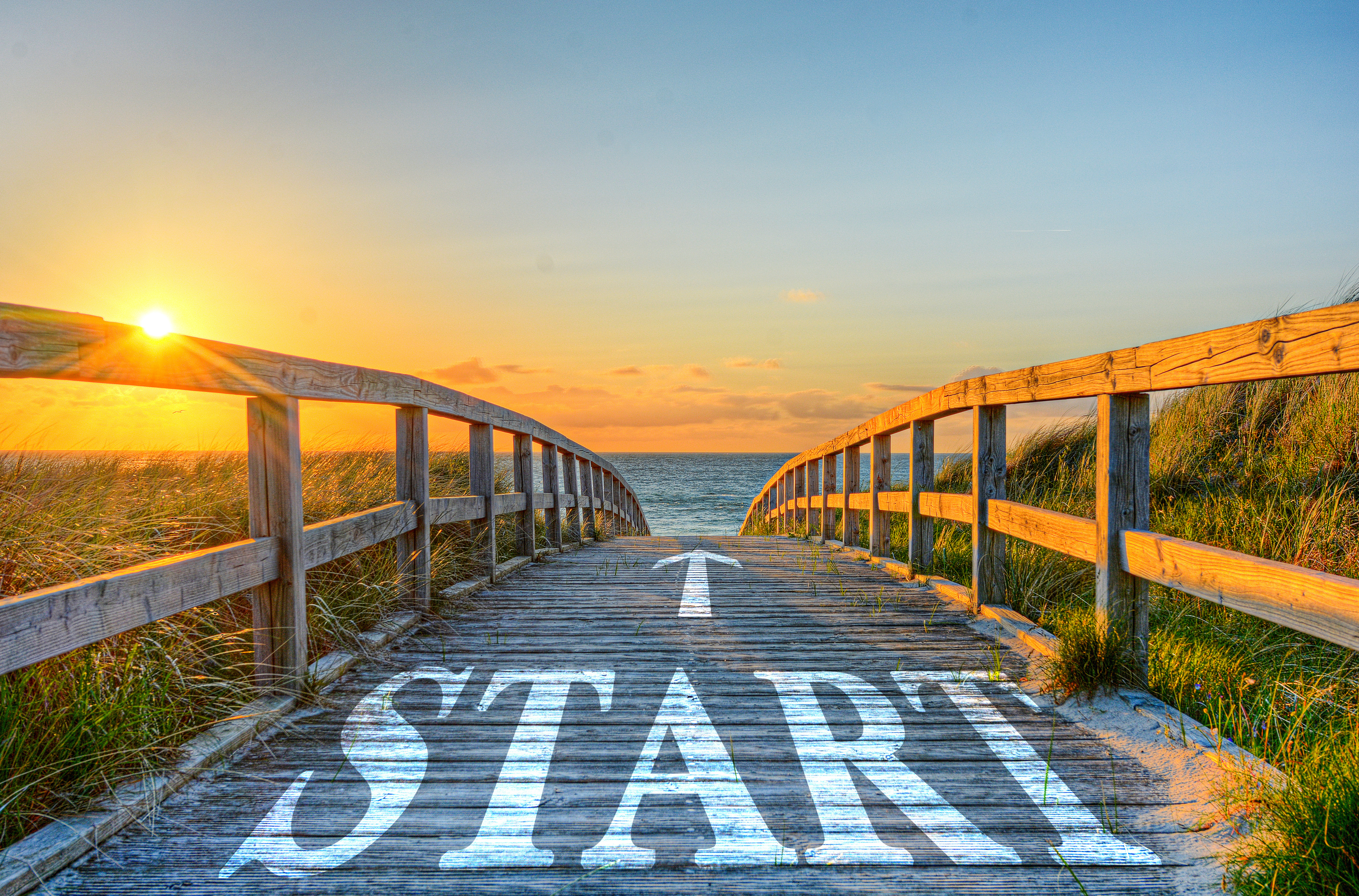 A wooden boardwalk with the word “START” painted on it leads toward the sunrise over the ocean, symbolizing the beginning of a writer’s journey with a book coach.