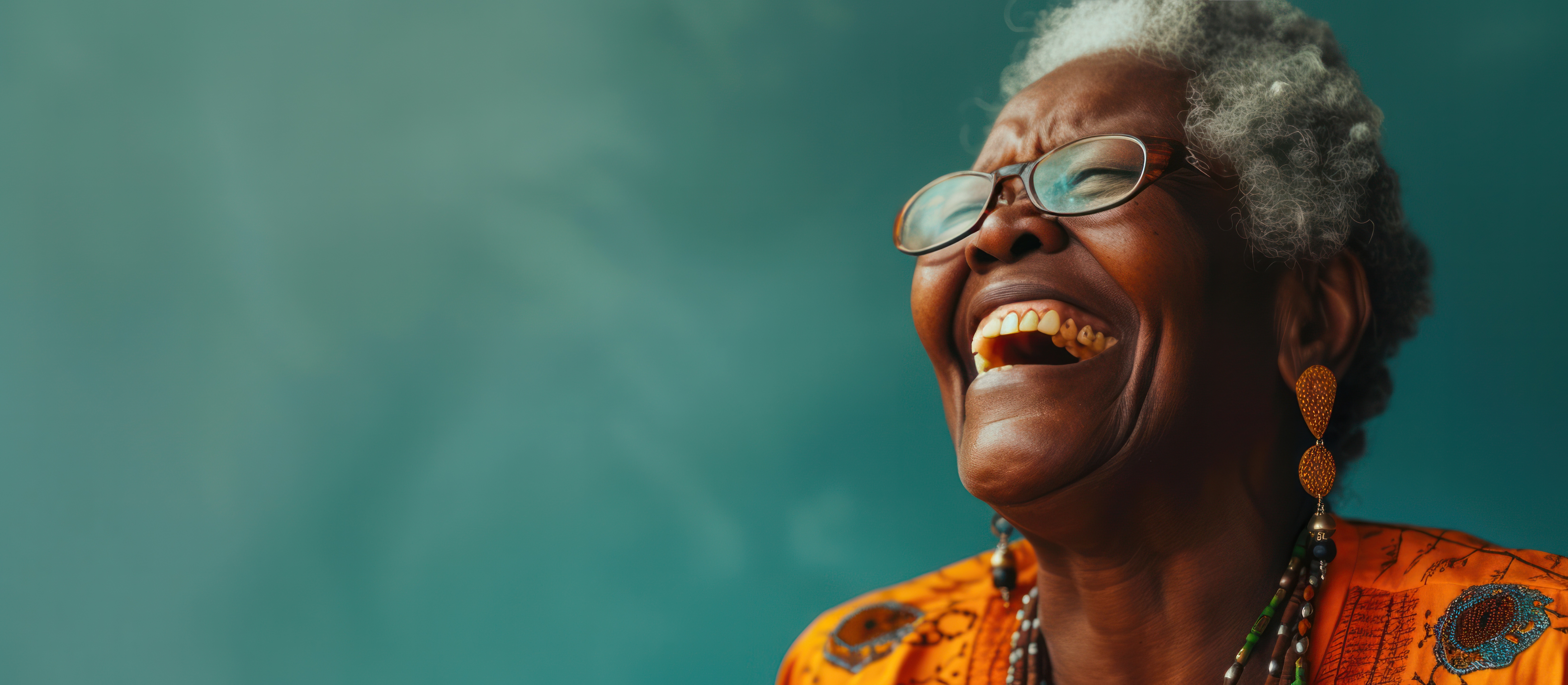 Smiling older woman with short gray hair and glasses laughing against a teal background, wearing an orange patterned top and beaded jewelry.